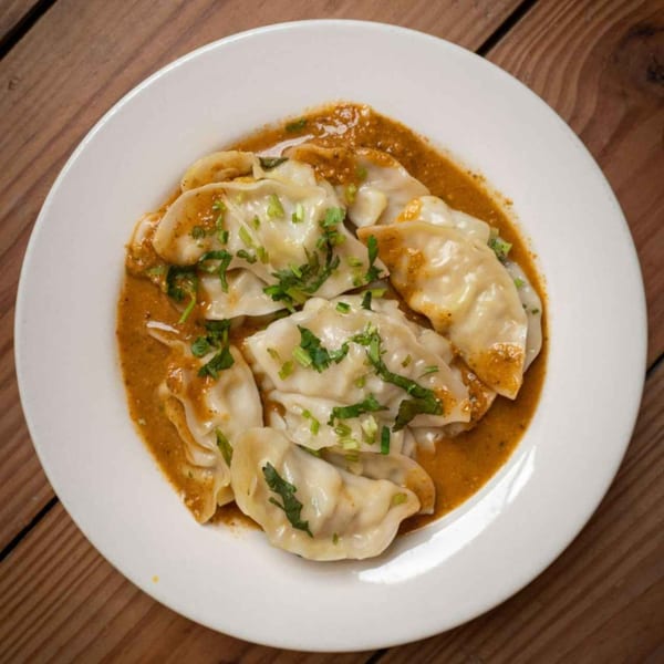 A white plate filled with Nepalese momo dumplings in a rich, orange sauce, garnished with fresh cilantro.