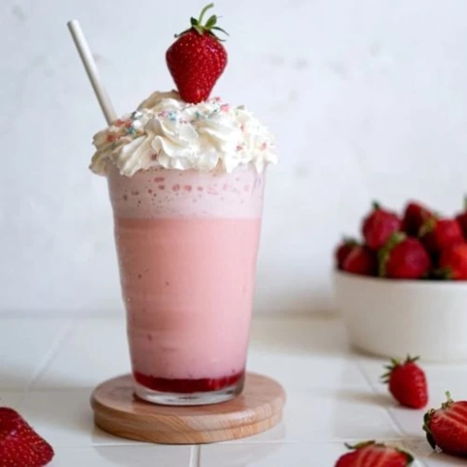 Pink strawberry milkshake with whipped cream, sprinkles, and a fresh strawberry on top.