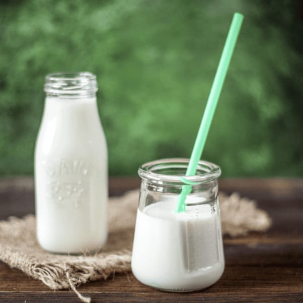 A glass bottle and a small jar of white lassi, with a green straw, on a wooden surface.