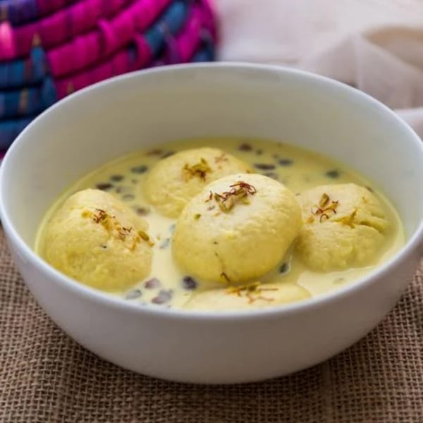 Four yellow Rasmalai dumplings in creamy milk, garnished with saffron threads, in a white bowl.