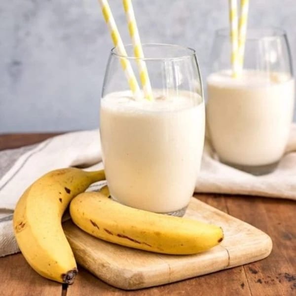 Two glasses of banana lassi with yellow striped straws, and two bananas on a wooden board.